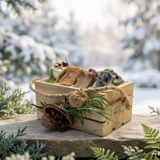 A small wooden gift box filled with assorted artisanal soaps, decorated with twine, a pinecone, and rosemary, set on a stone surface with a snowy outdoor background.