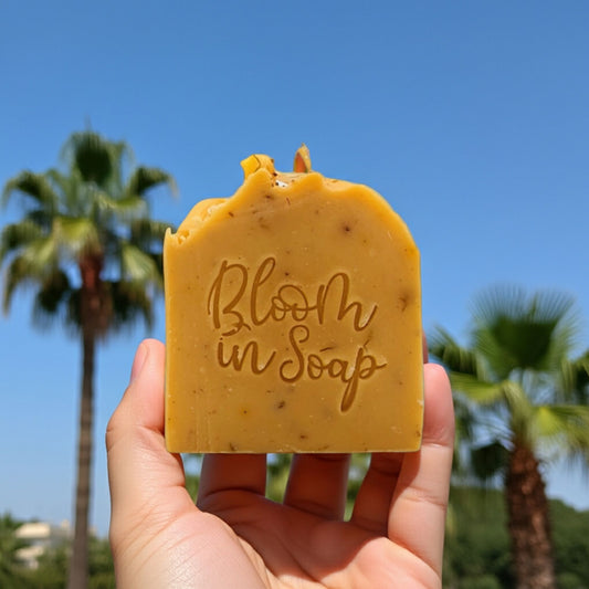 Bar of soap with 'Bloom in Soap' text held in front of palm trees