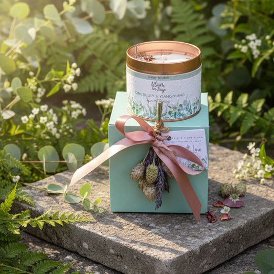 Candle in a decorative box with a pink ribbon on a stone surface with greenery in the background
