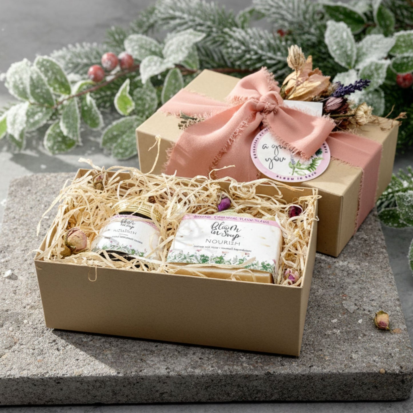 Gift set with soaps and a jar, in a box wrapped with a pink ribbon, on a stone surface with greenery in the background.