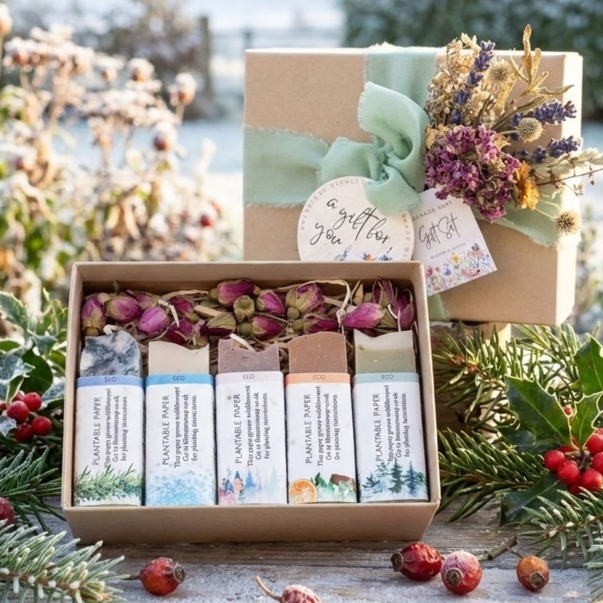 A gift set of five handcrafted soap bars in a kraft box, displayed alongside a matching wrapped gift box with a mint ribbon and dried flowers. The soaps feature winter-themed illustrations and are surrounded by festive greenery, holly, and red berries on a rustic wooden surface.