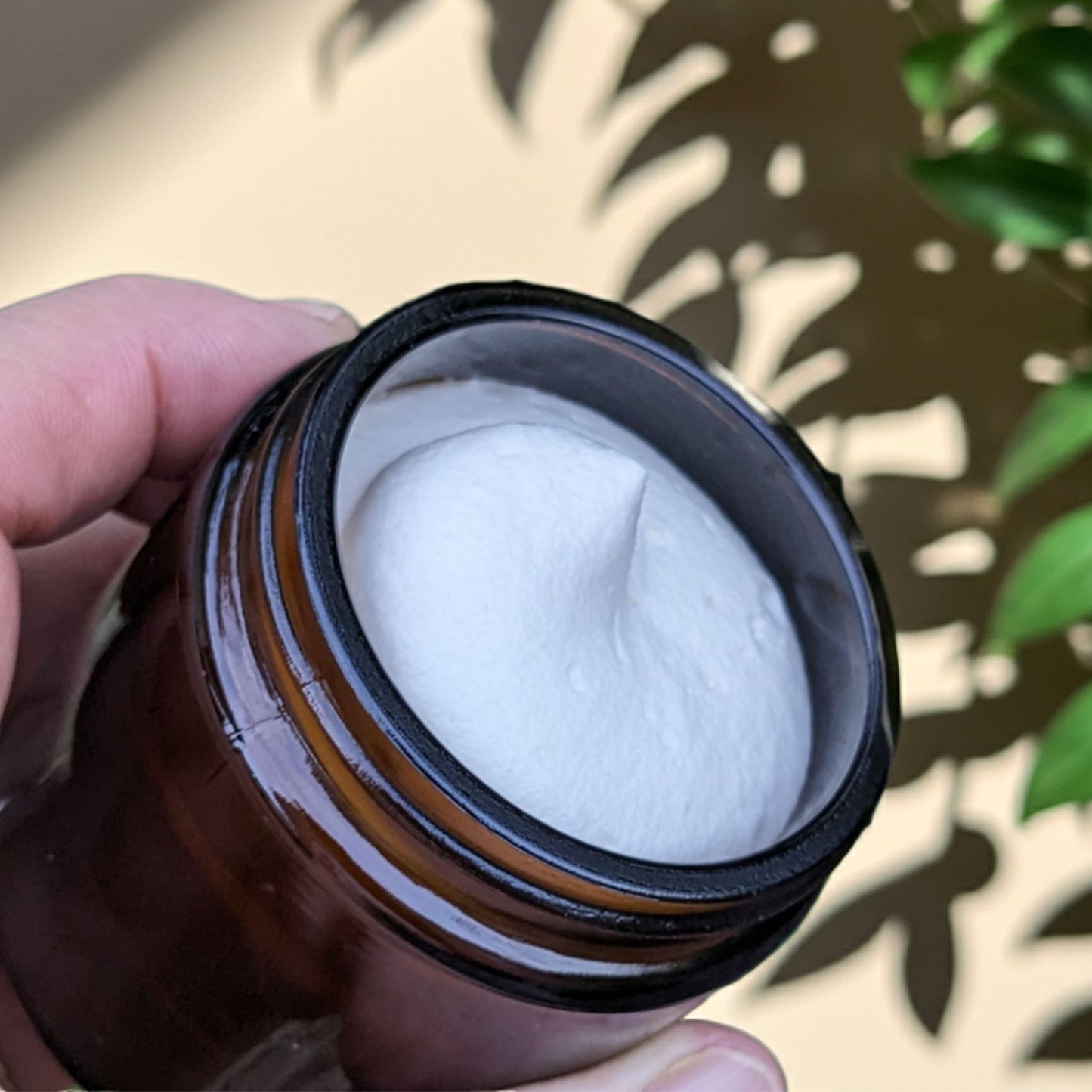Brown jar with white cream inside, held by a hand against a leafy background