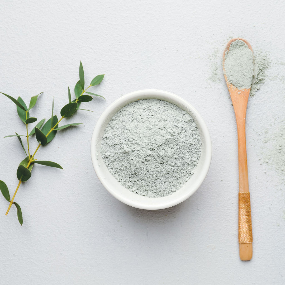 A white ceramic bowl filled with light gray-green powder, styled on a white surface with a wooden spoon holding more of the powder and a small green leaf sprig.