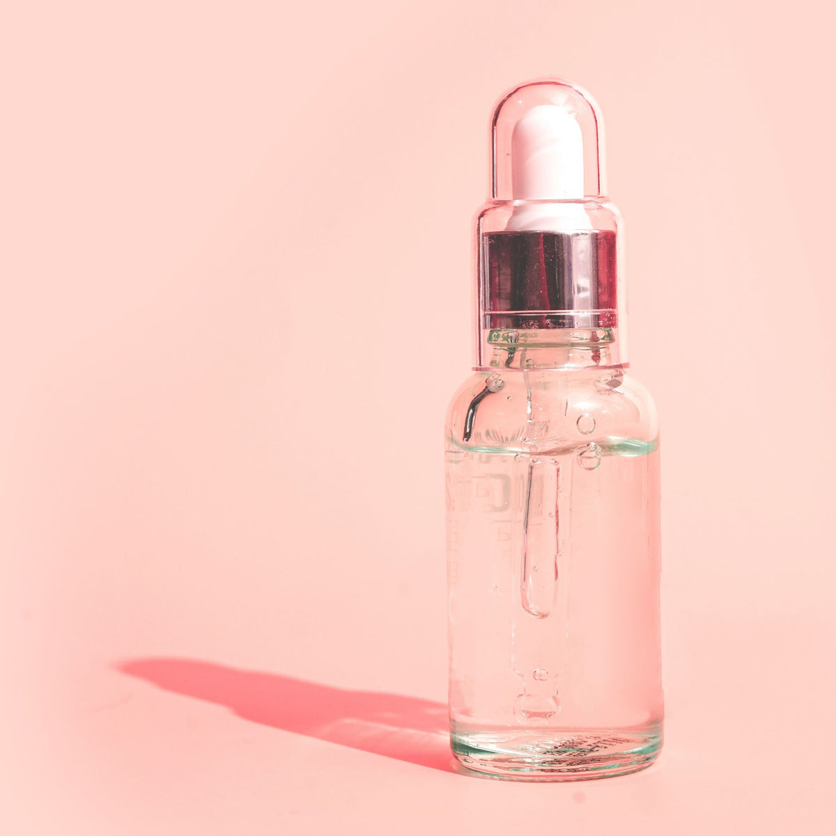 A clear glass dropper bottle filled with a transparent liquid, featuring a metallic silver collar and a white rubber bulb, set against a solid pink background with a soft pink shadow.
