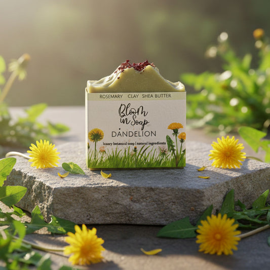 Bar of soap with 'Bloom in Soap' packaging on a stone surface surrounded by dandelions.