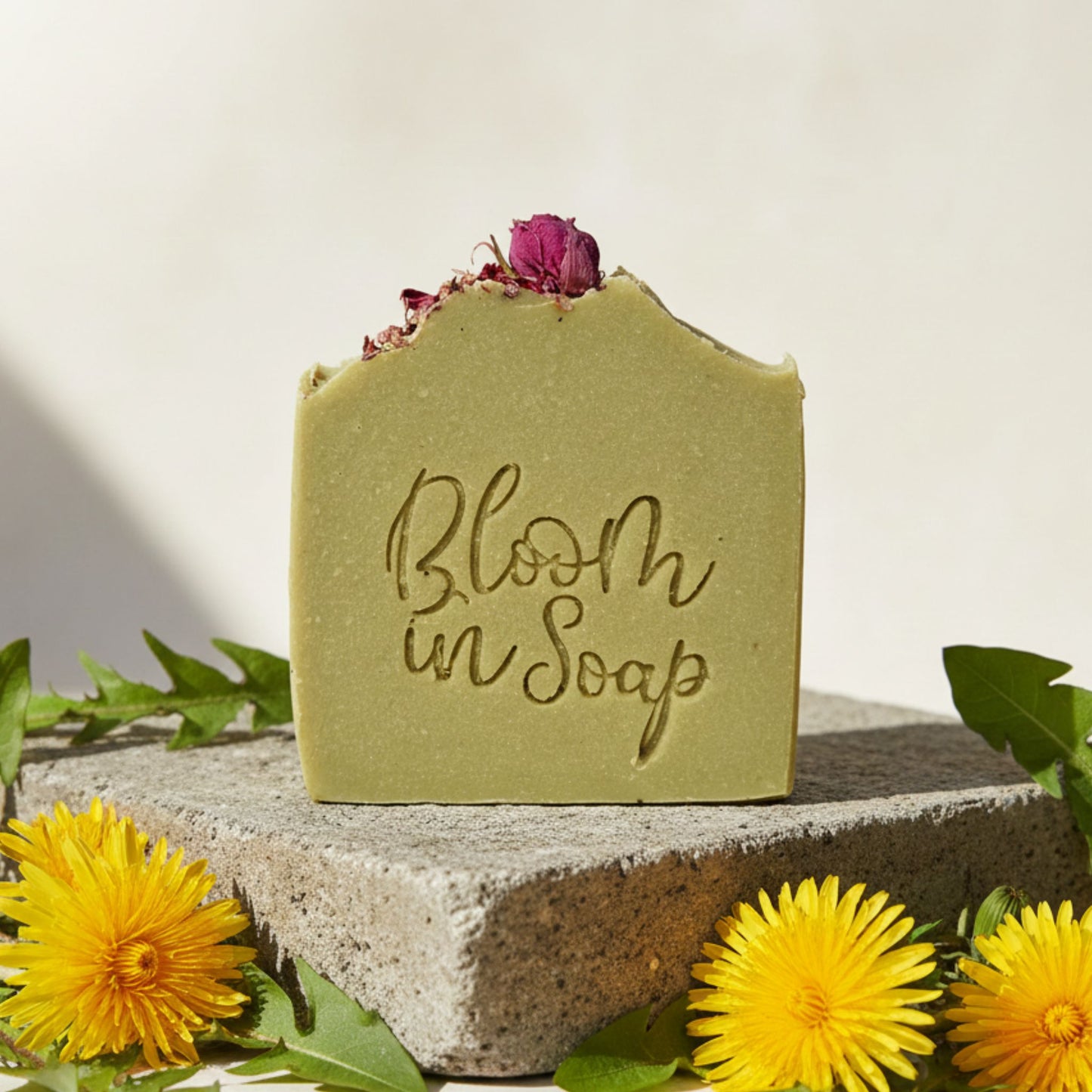 Bar of soap with 'Bloom in Soap' text, surrounded by yellow flowers and green leaves on a light background