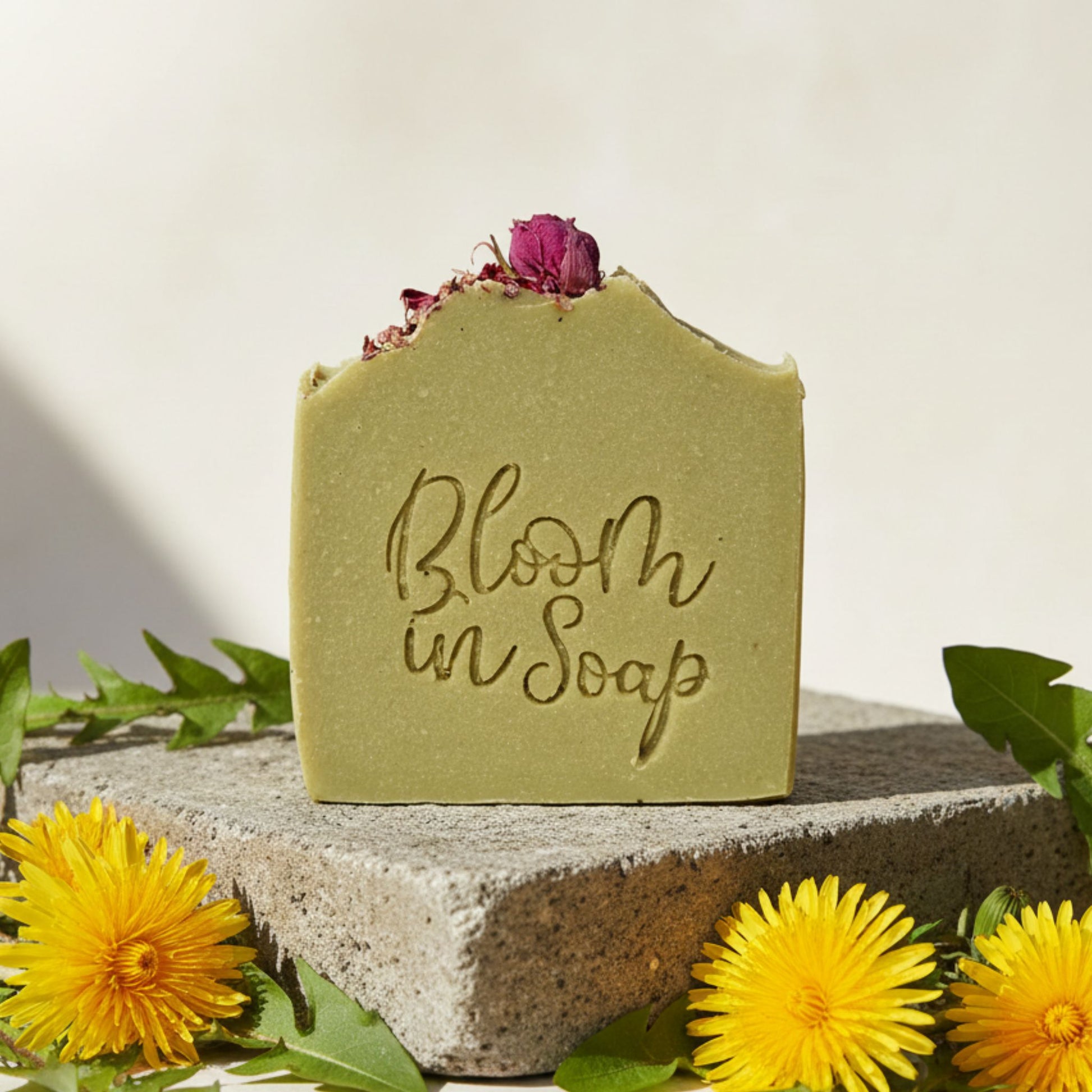 Bar of soap with 'Bloom in Soap' text, surrounded by yellow flowers and green leaves on a light background