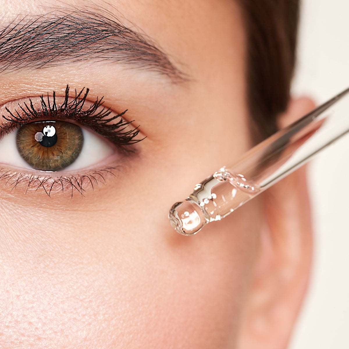A close-up of a person's eye with a glass dropper positioned near the eye area, showing a clear liquid being applied. The dropper contains transparent serum or oil.