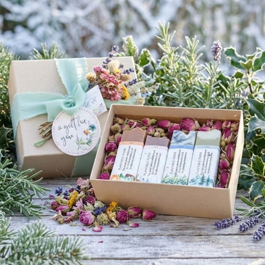 A gift set of natural skincare products in a kraft paper box, displayed next to its matching gift box on a rustic wooden surface surrounded by greenery and dried flowers. The set includes four handmade soap bars.