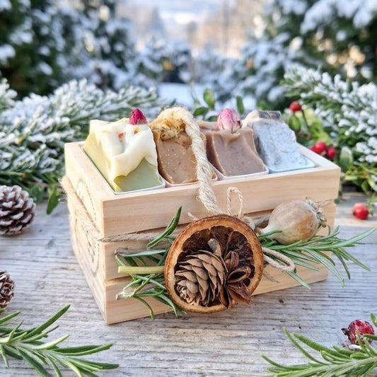 A wooden gift box containing four artisan soap bars, styled with rosemary, a dried orange slice, pinecones, and red berries on a rustic wooden table with a snowy outdoor background.