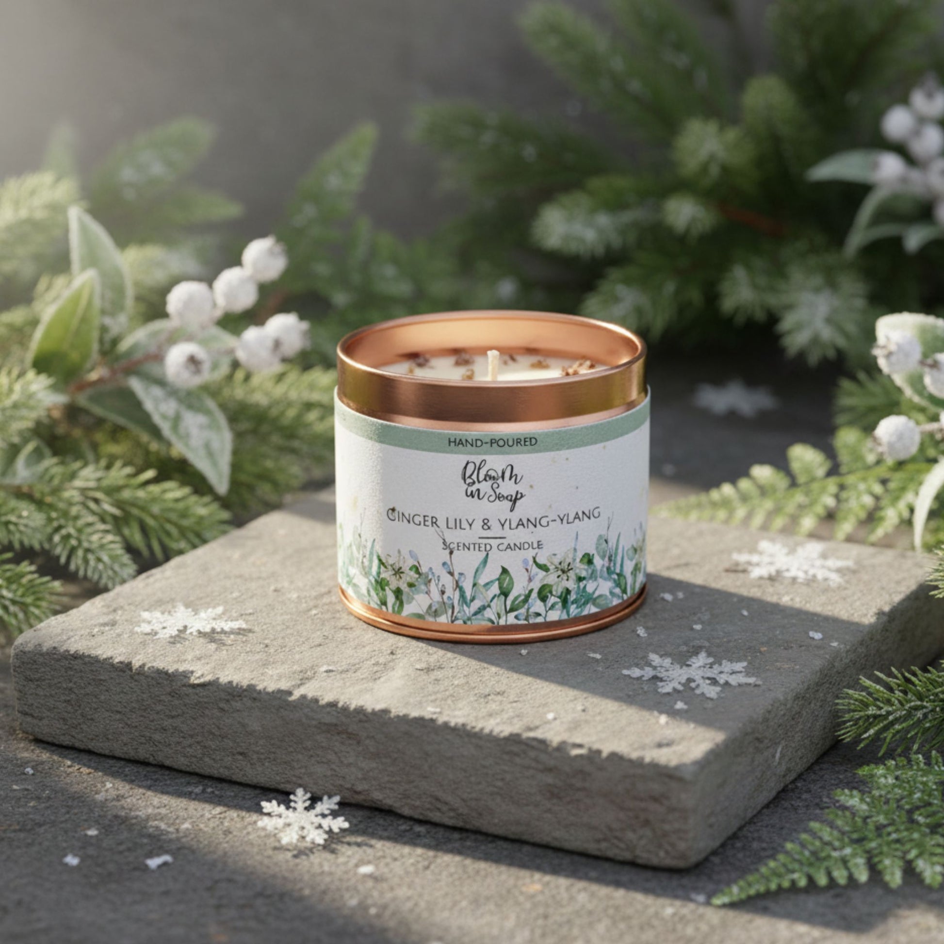 Candle in a decorative tin with a green label on a stone surface with snowflakes and greenery.