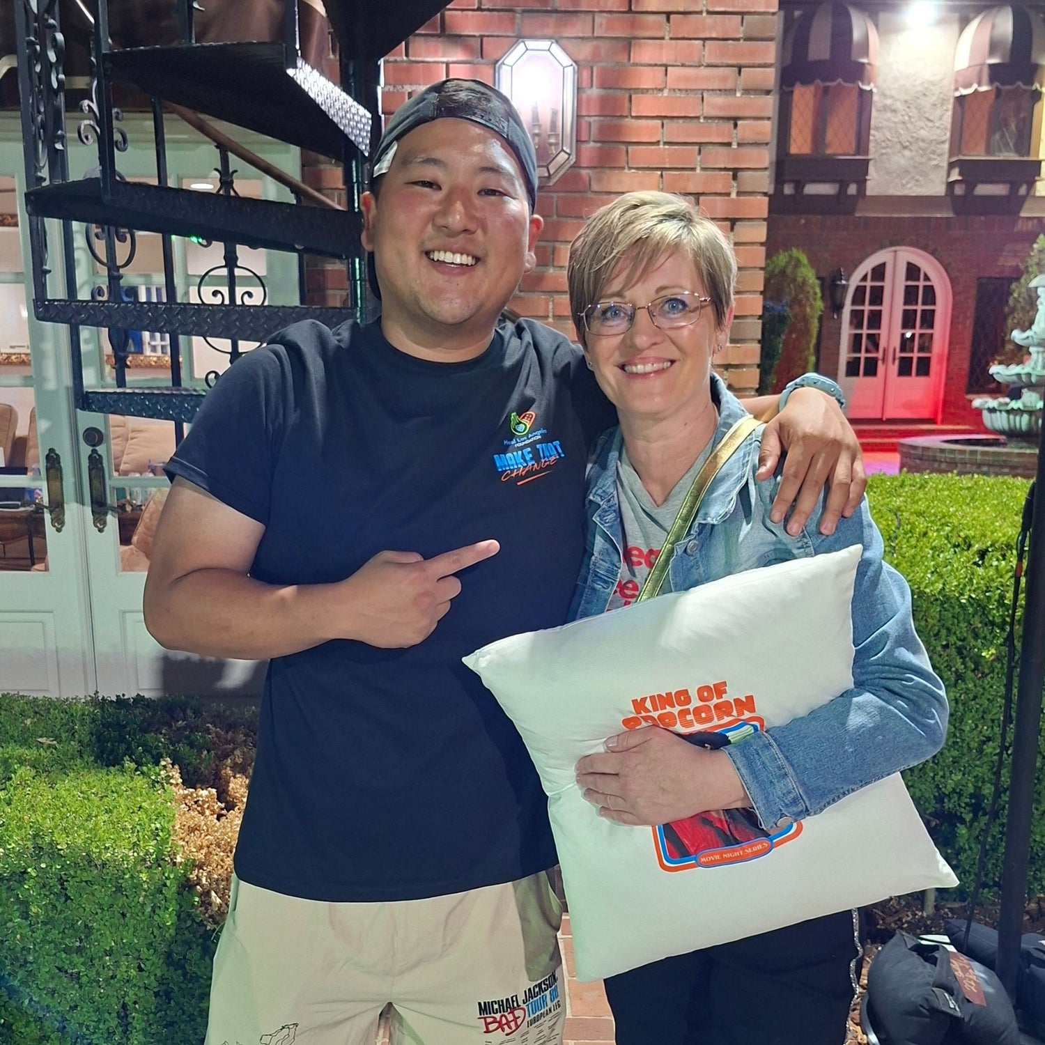 Two people standing outdoors with a decorative house in the background, one holding a pillow with text.