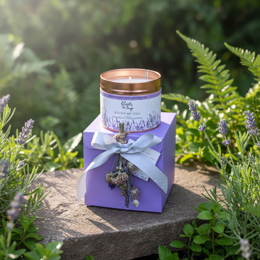 Lavender-scented candle in a purple box with lavender flowers on a stone surface surrounded by greenery.