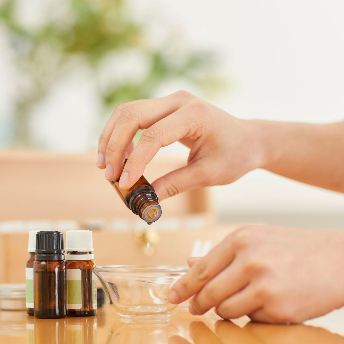 A close-up of a person's hands holding an amber glass essential oil bottle and pouring a drop of liquid into a small clear glass bowl, with two more amber bottles in the background on a wooden table.