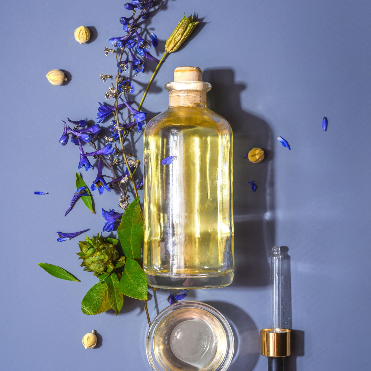 A clear glass bottle filled with pale yellow oil, styled on a lavender background with blue flowers, green leaves, small beige pods, a small clear jar, and a glass dropper with a gold collar.