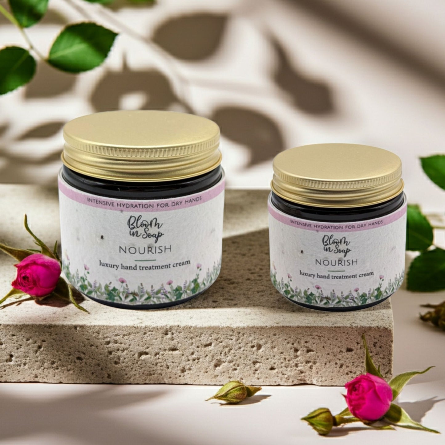 Two jars of 'Nourish' hand treatment cream with gold lids on a stone surface with pink flowers and green leaves.