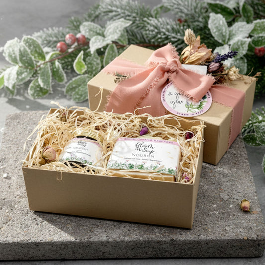 Gift set with soaps and a jar, in a box wrapped with a pink ribbon, on a stone surface with greenery in the background.