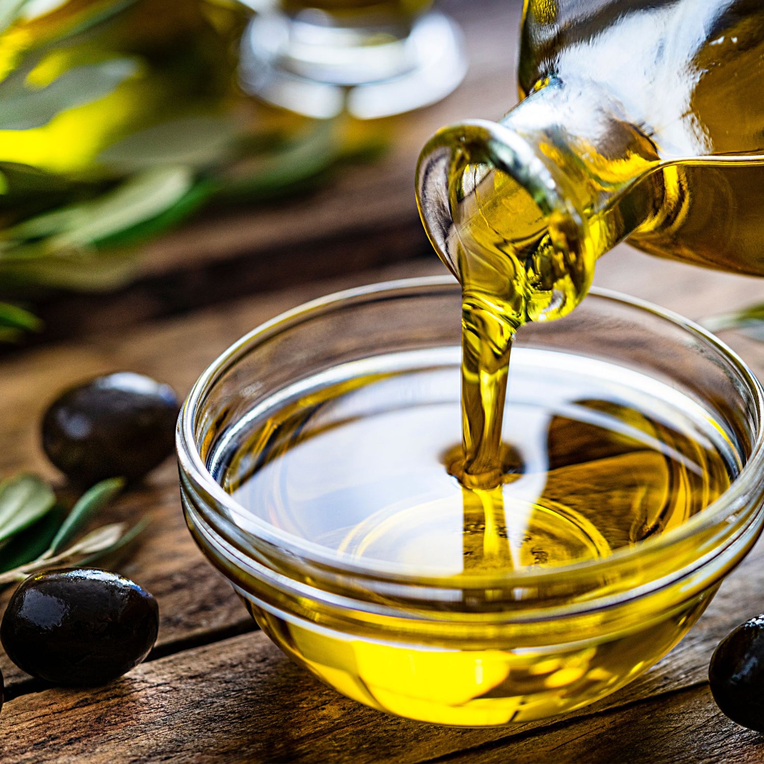 Olive oil being poured into a glass bowl with olives and olive branches on a wooden surface.