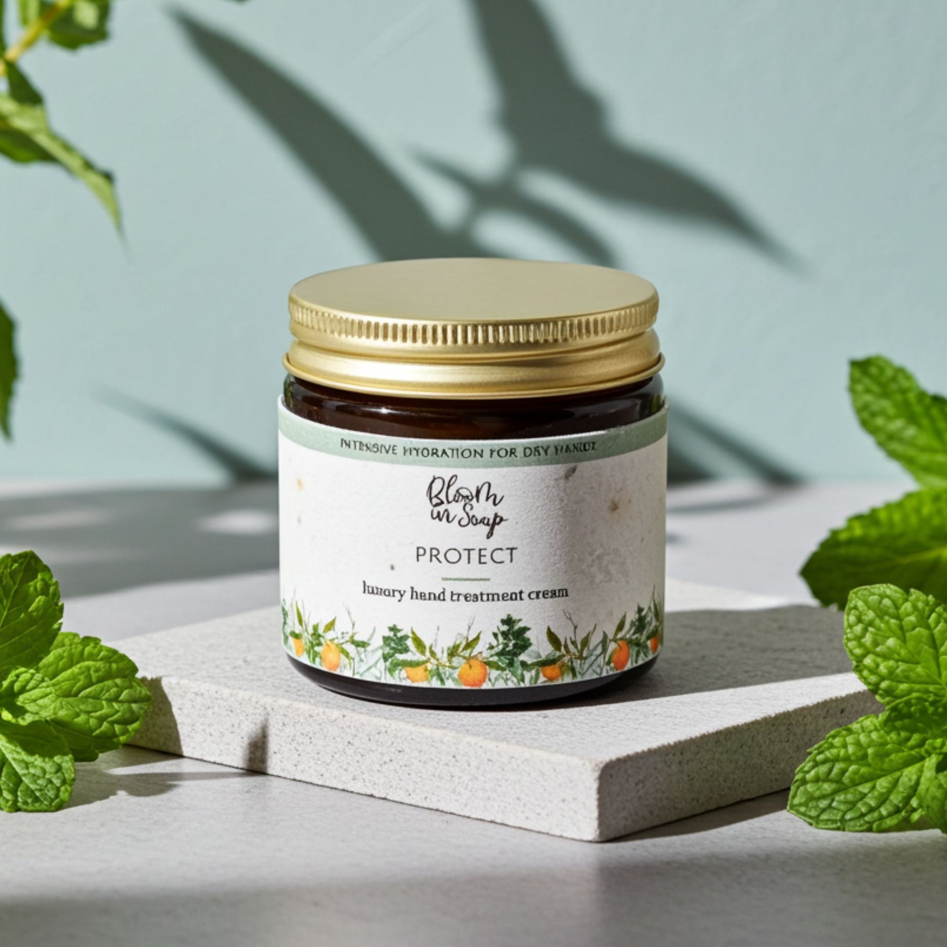 Jar of hand treatment cream with a gold lid on a light surface surrounded by green leaves.