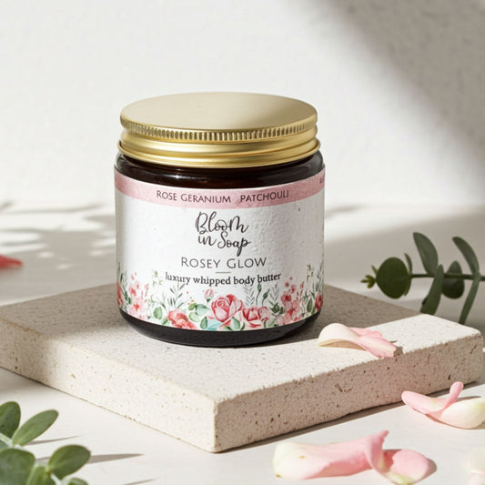 Jar of 'Rosey Glow' body butter with floral design on label, placed on a light surface with pink petals and green leaves.