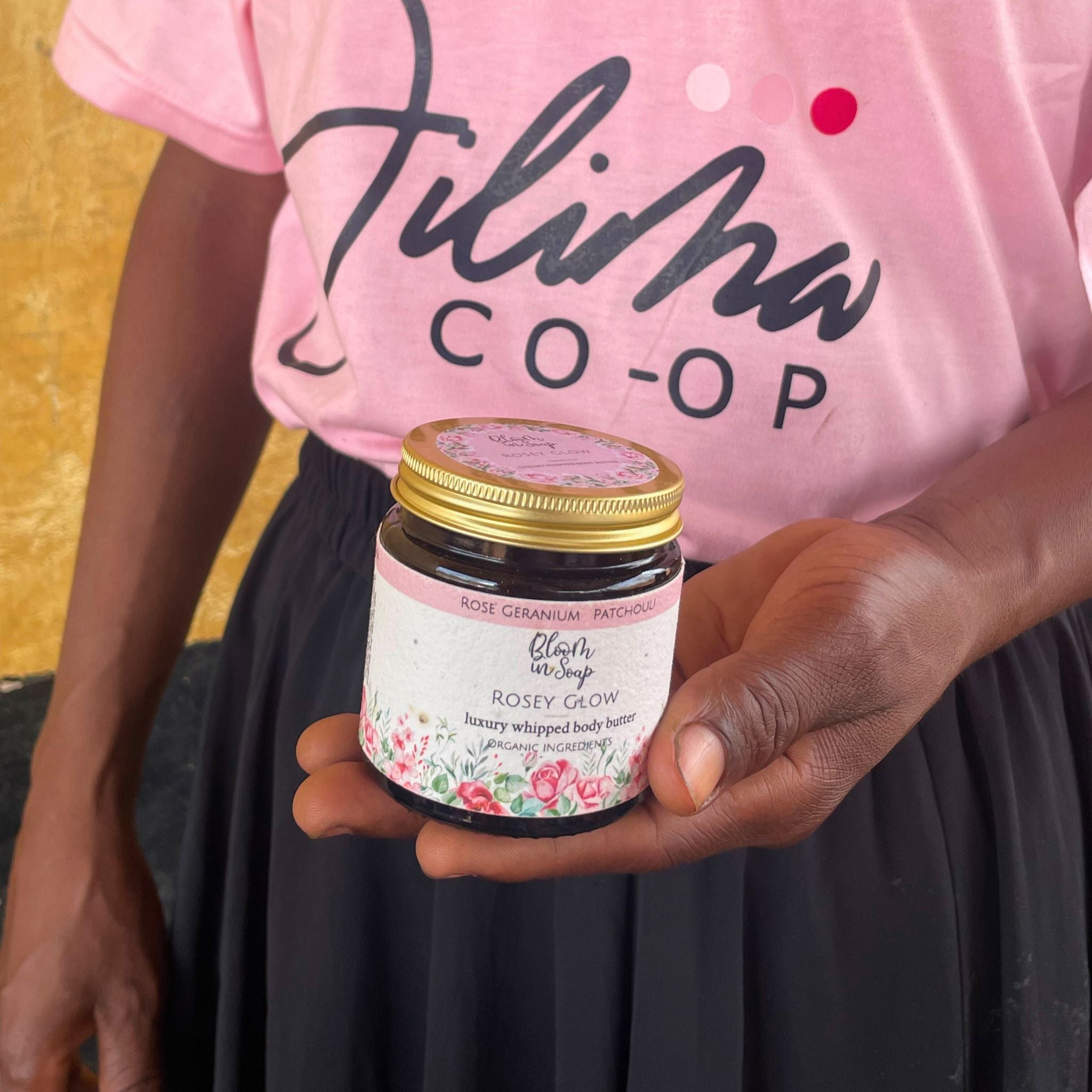 Woman wearing a pink shirt with 'Jilima Co-op' holding a jar labeled 'Rosey Glow'.
