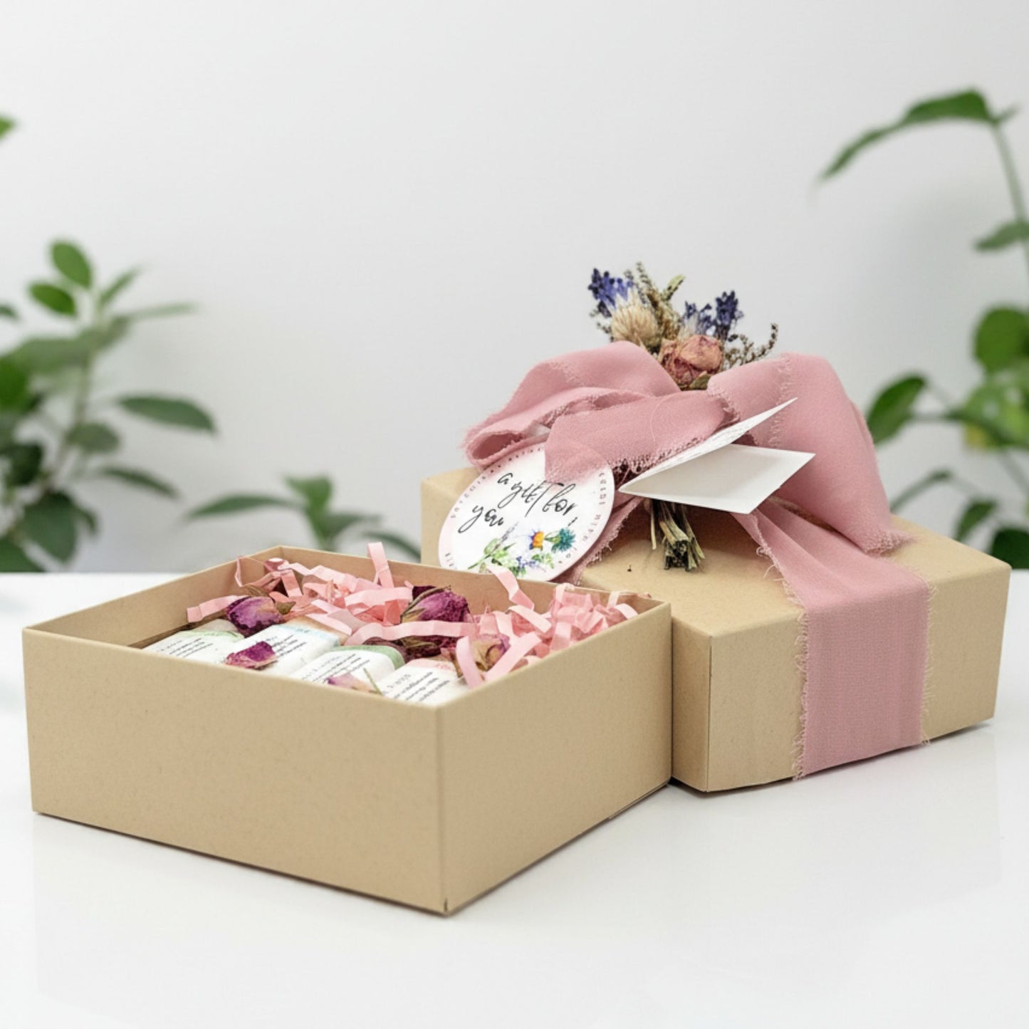 Gift box with pink ribbon, flowers, and a card on a white surface with green plants in the background.
